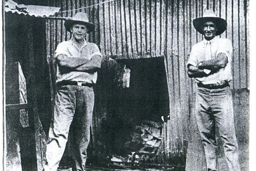 A black and white image of two men standing with arms crossed and hats on beside a blown-up wall.