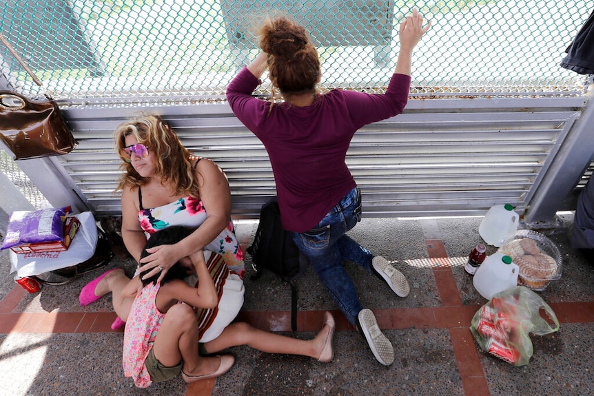 A woman cuddles a young girl while an older girl looks through a wire fence, their belongings surrounding them on the ground