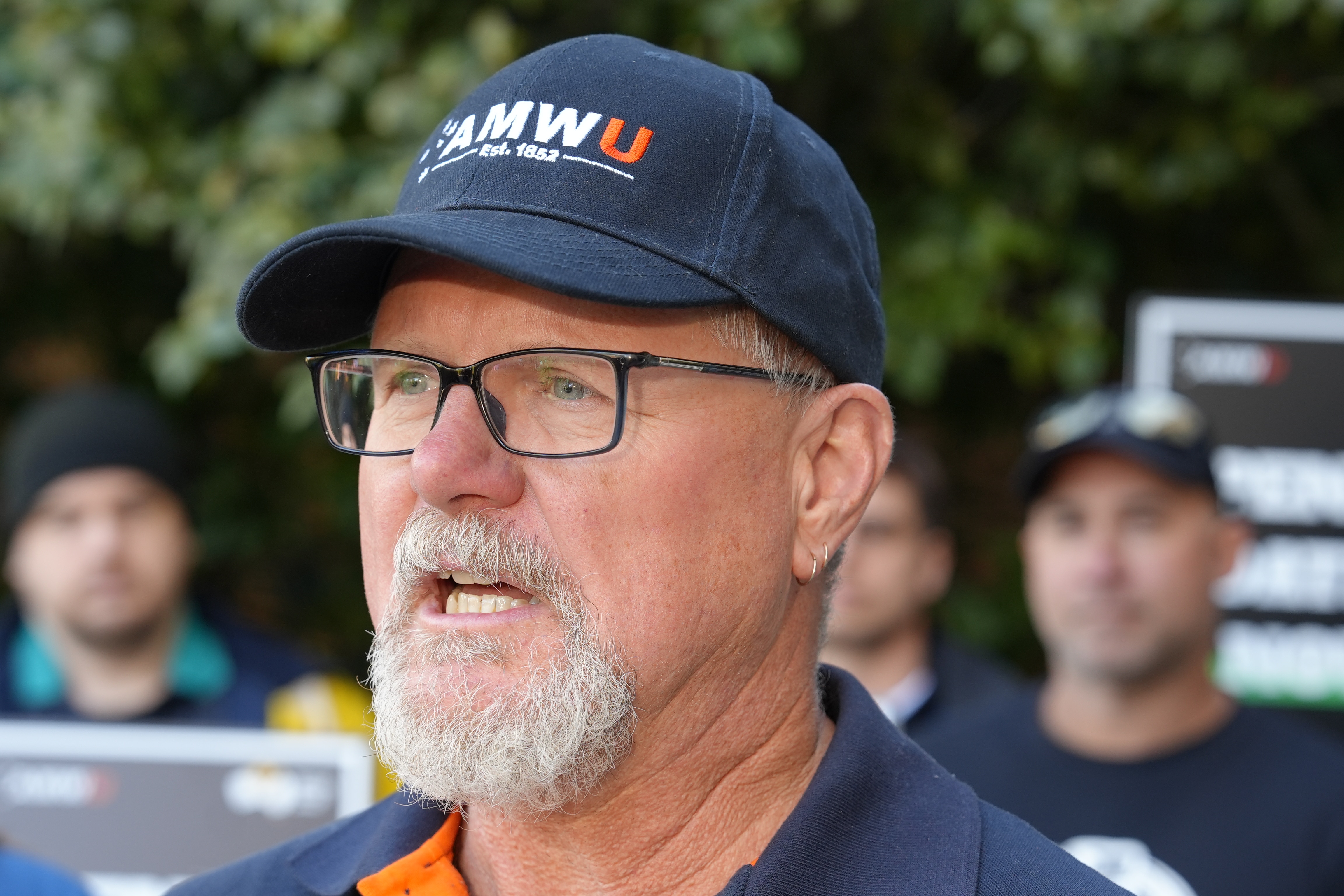 A man with a short grey goatie, dark square glasses and a navy cap with AMWU on the front.