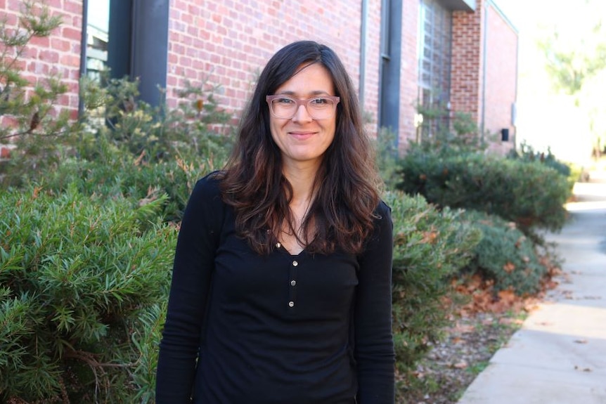 A woman with long brown hair and glasses stands in front of a red brick building wearing a black shirt.