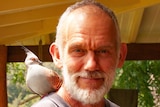 A balding man with a grey beard with a dove sitting on his shoulder, smiles at the camera.