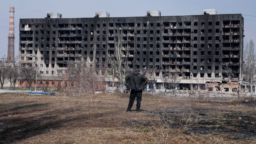 A man with a walking stick looks at a burned apartment building from a barren patch of land some distance away.