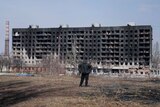 A man with a walking stick looks at a burned apartment building from a barren patch of land some distance away.