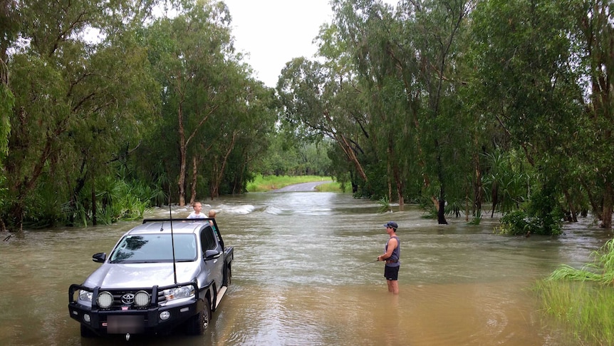 A man stands in the water fishing, while two young boys stand in the back of a ute.