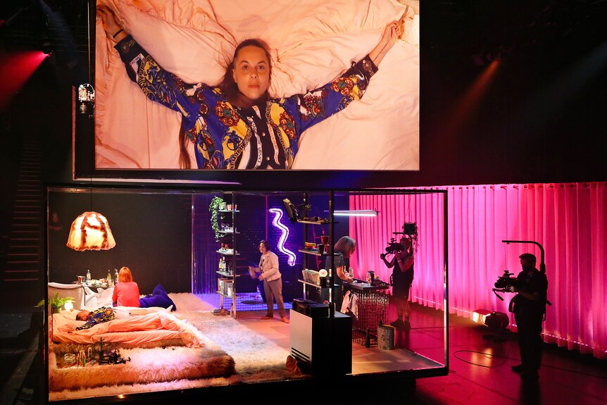One woman lies on a bed, arms outstretched, a close-up of her seen on a hanging screen. Three other women are on stage with her.