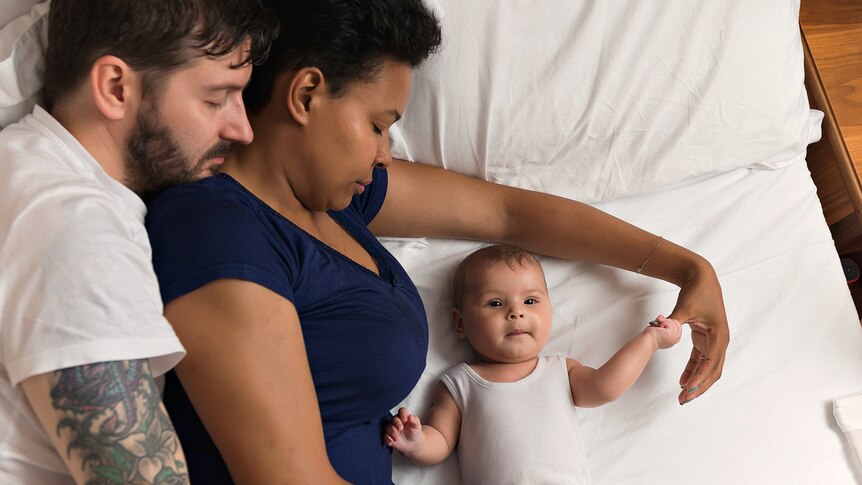 Two parents lying in a bed asleep with an awake baby next to them