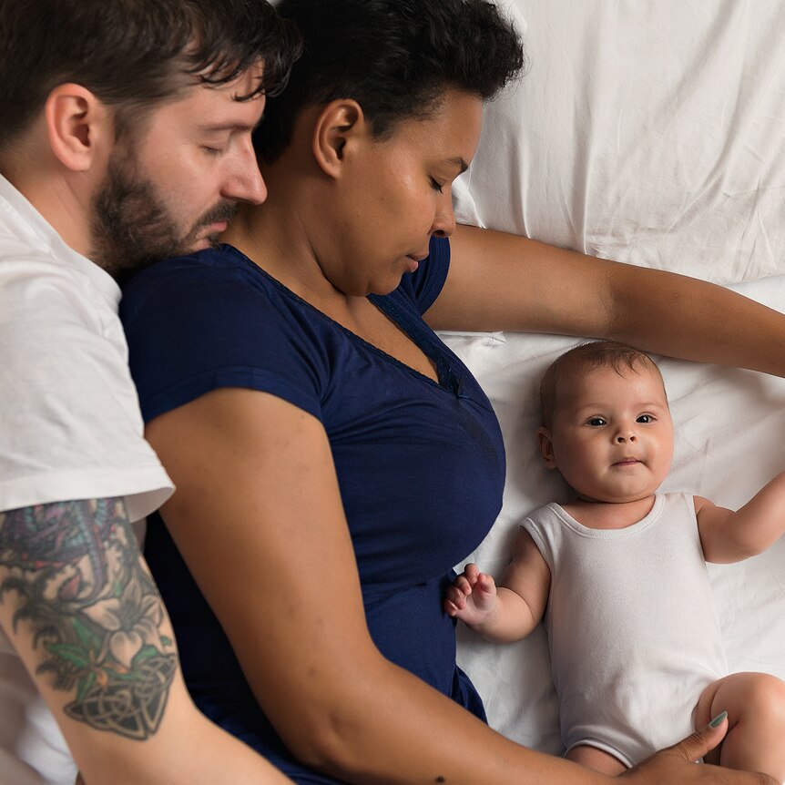 Two parents lying in a bed asleep with an awake baby next to them