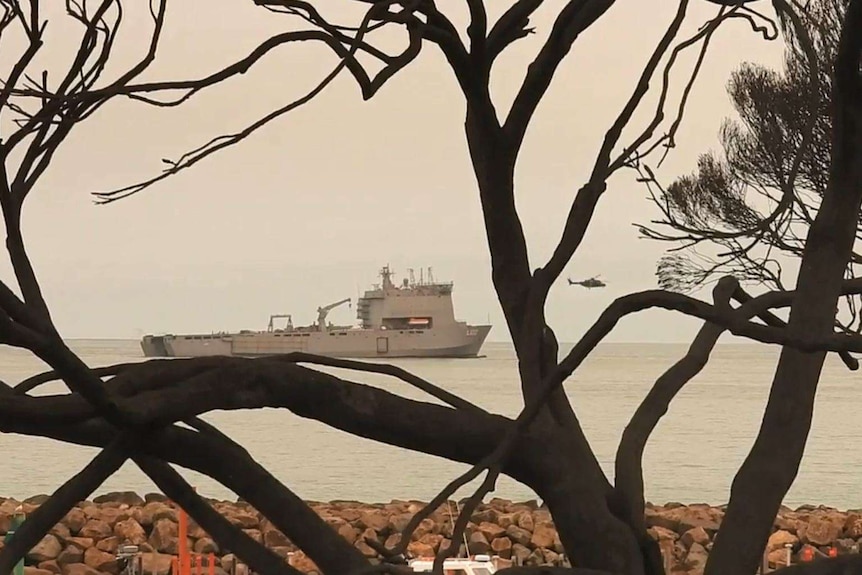 A helicopter leaves a large navy ship in smoke-covered water.