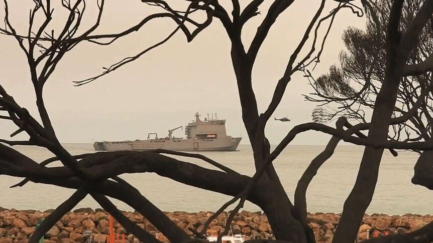 A helicopter leaves a large navy ship in smoke-covered water.