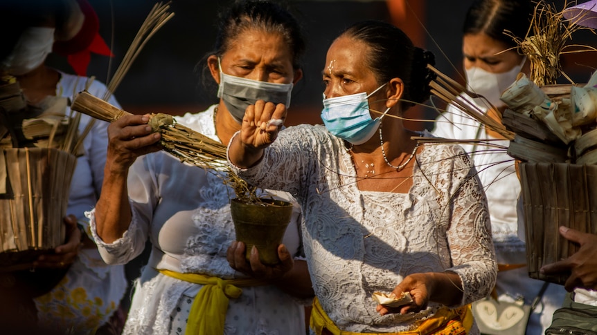 Two women hold sage and sprinkle holy water while wearing face masks and white dresses.