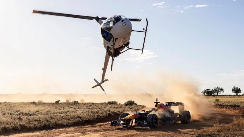 a low-flying helicopter passes over a F1 racing car on an outback road