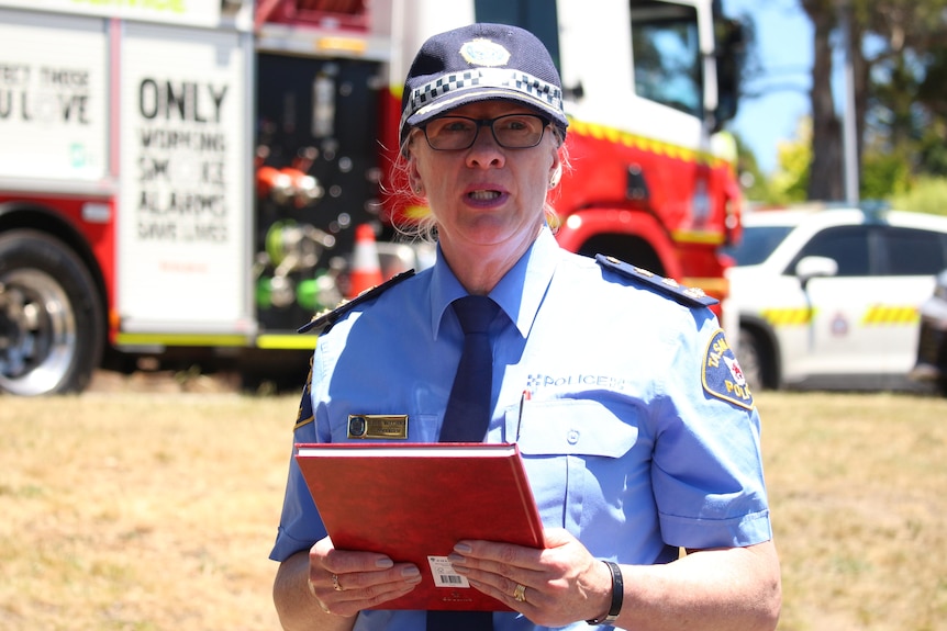 A police officer holds a notebook standing in front of emergency service vehicles.