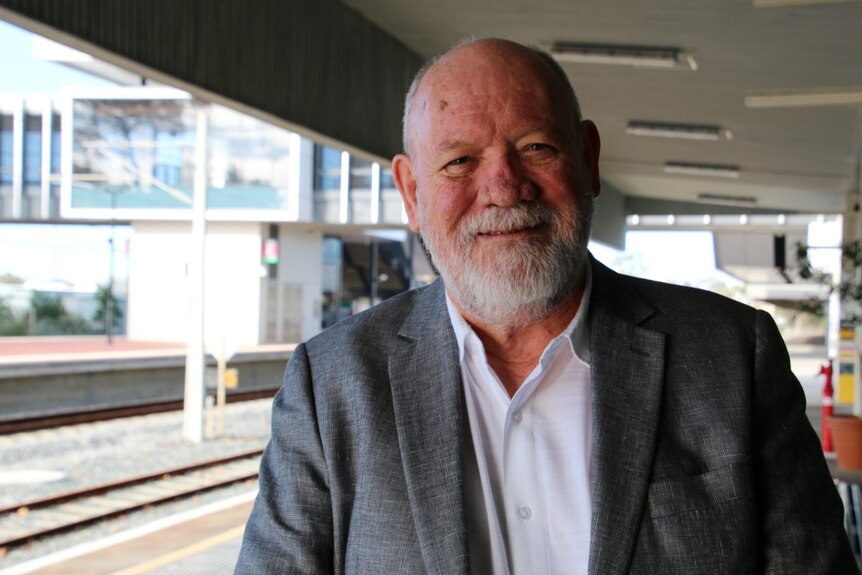 A man wearing an open-neck white shirt and grey blazer stands on a train platform.