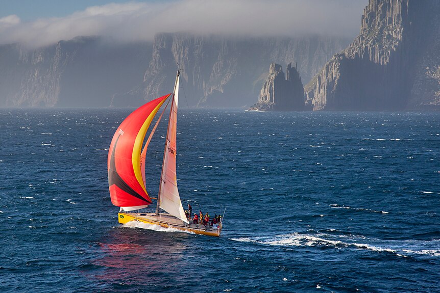 A yacht with a red black and yellow sail sails past cliffs 
