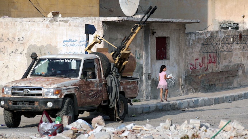A young girl walks past a truck parked on a road strewn with rubble.