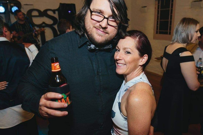 Anita and Mitch smile at a party. Mitch has one arm around Anita and with the other arm raises a beer.
