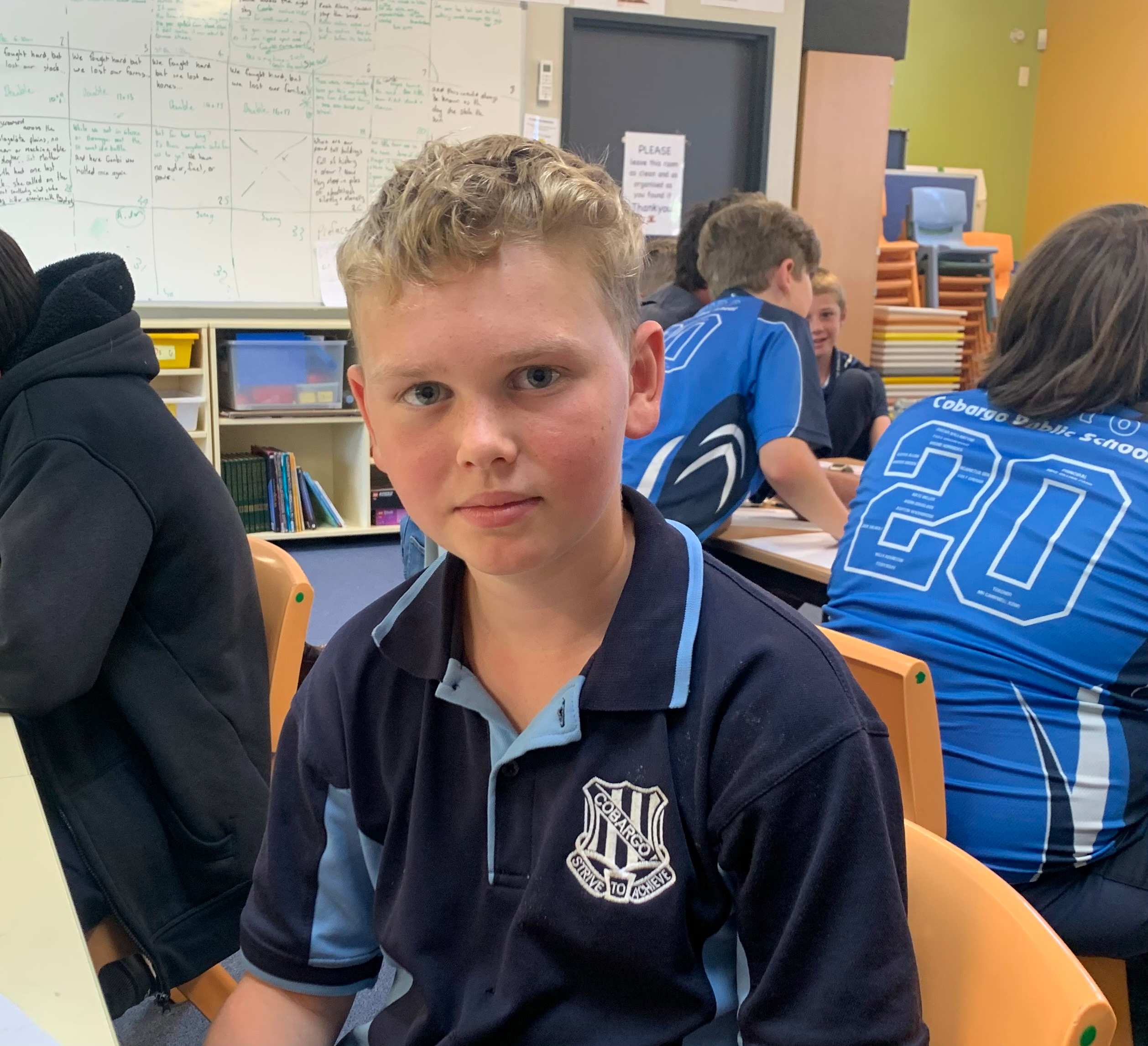 A boy in a blue school uniform sitting in a classroom with a white board and other students behind him