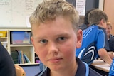 A boy in a blue school uniform sitting in a classroom with a white board and other students behind him