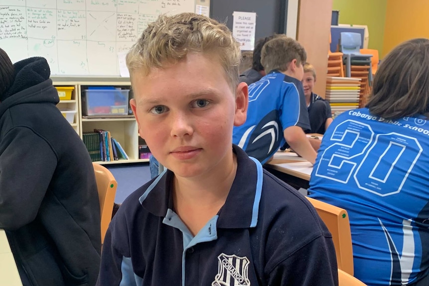 A boy in a blue school uniform sitting in a classroom with a white board and other students behind him