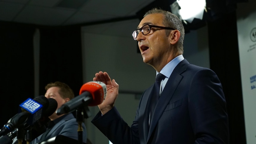 A man wearing a suit and tie speaks in front of microphones