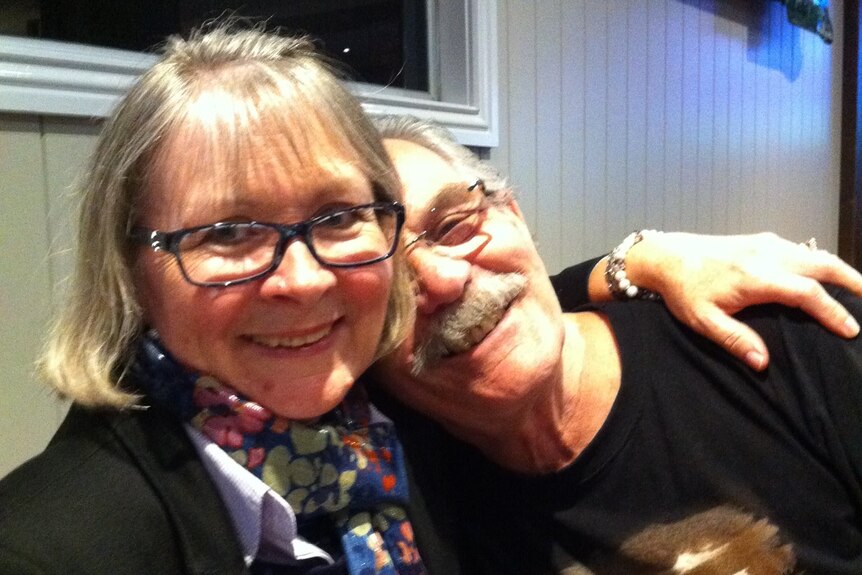 A smiling woman with her grey moustached husband resting his head on her shoulder.