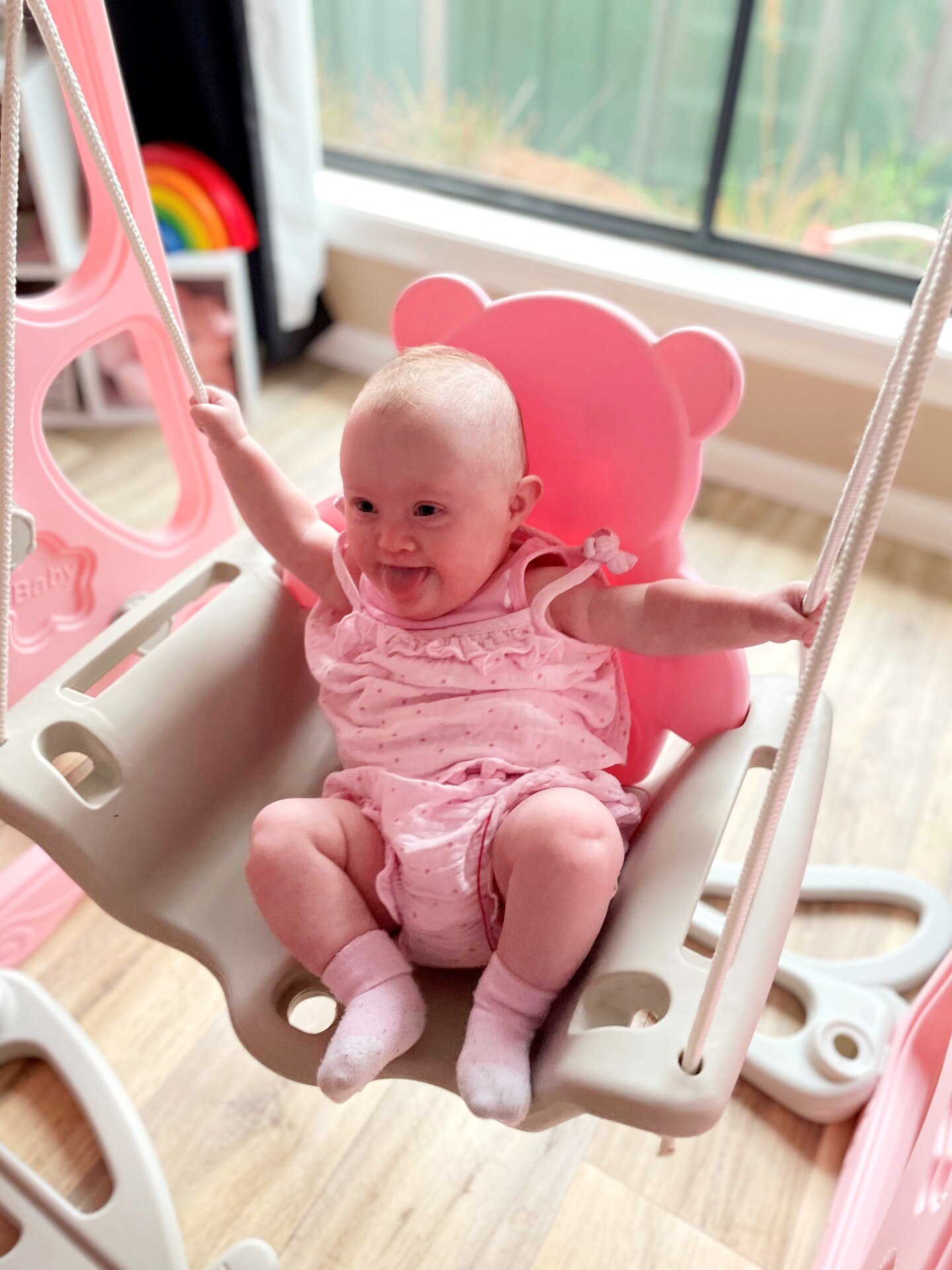 Baby Arlee is wearing a pink jumpsuit and sitting on a little bear-shaped swing. She is smiling and poking her tongue out.