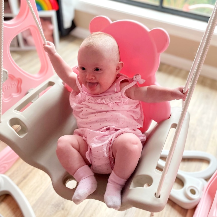 Baby Arlee is wearing a pink jumpsuit and sitting on a little bear-shaped swing. She is smiling and poking her tongue out.