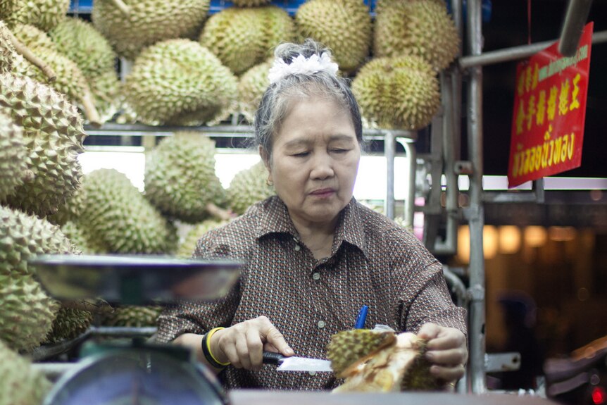 A woman with black grey top knot cuts up durian with knife in front and next to a wall of durian at night time in market stall.