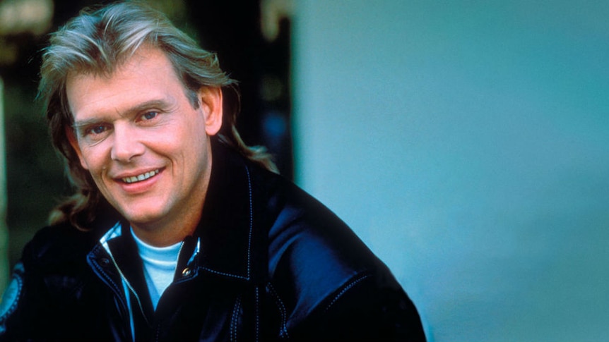 A young John Farnham with long hair, smiling and wearing a leather jacket.