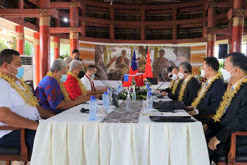 Eight men in formal wear, masks and leis sit around a table in an open-room with heavy poles holding up the roof