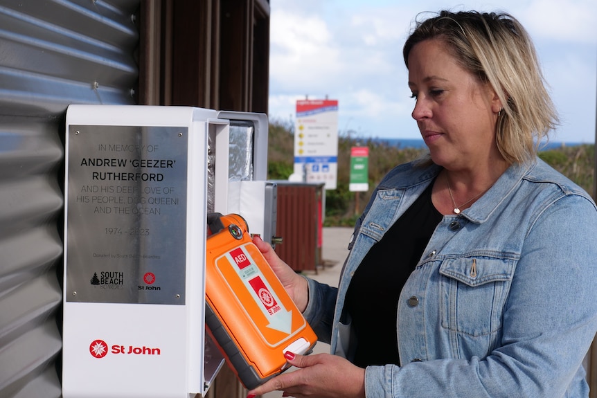 Renee holding a defibrillator next to its box. The box has a plaque to Andrew 'Geezer' Rutherford.