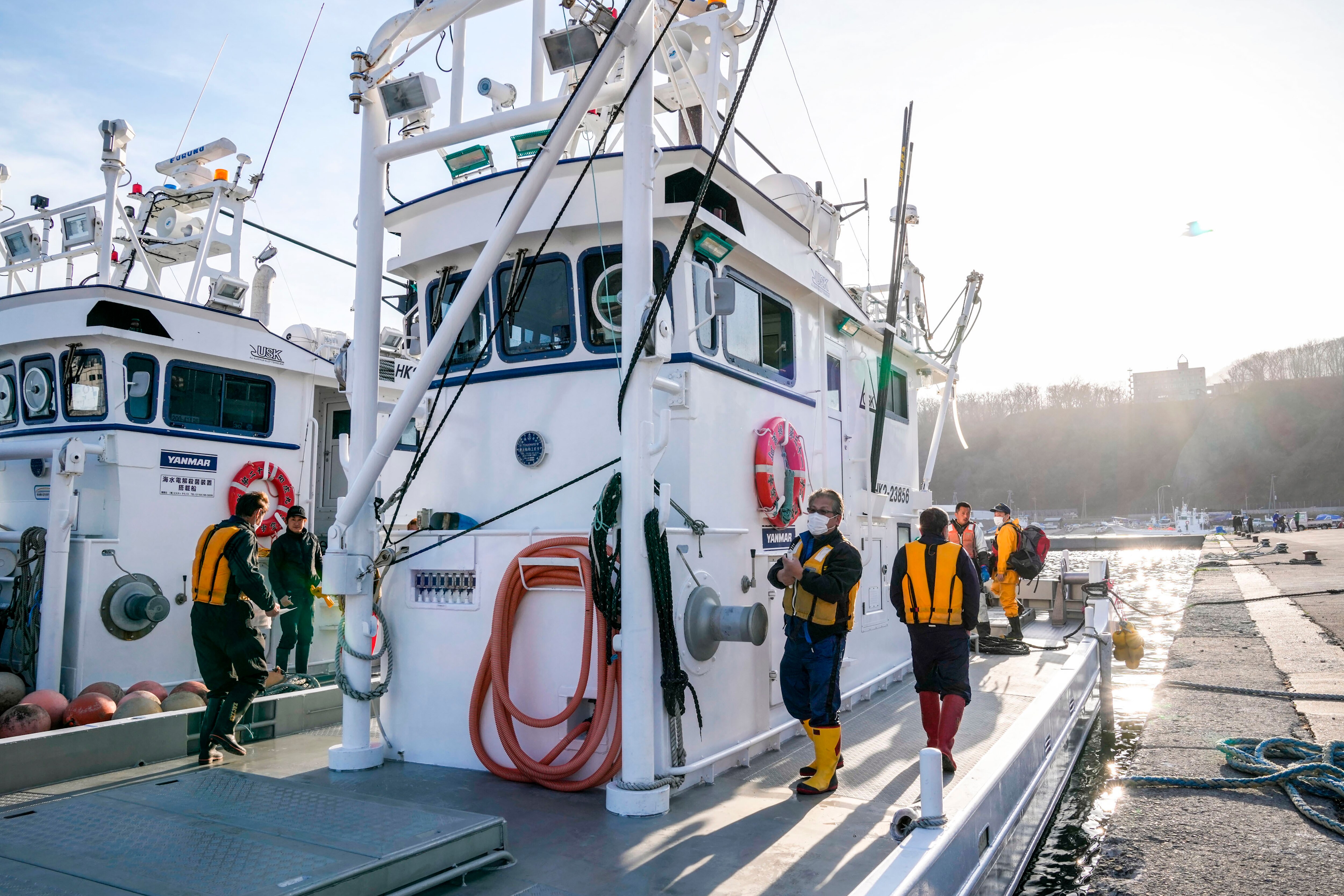 Crew members of a fishing boat stand on its deck, wearing high-viz jackets and high boots.