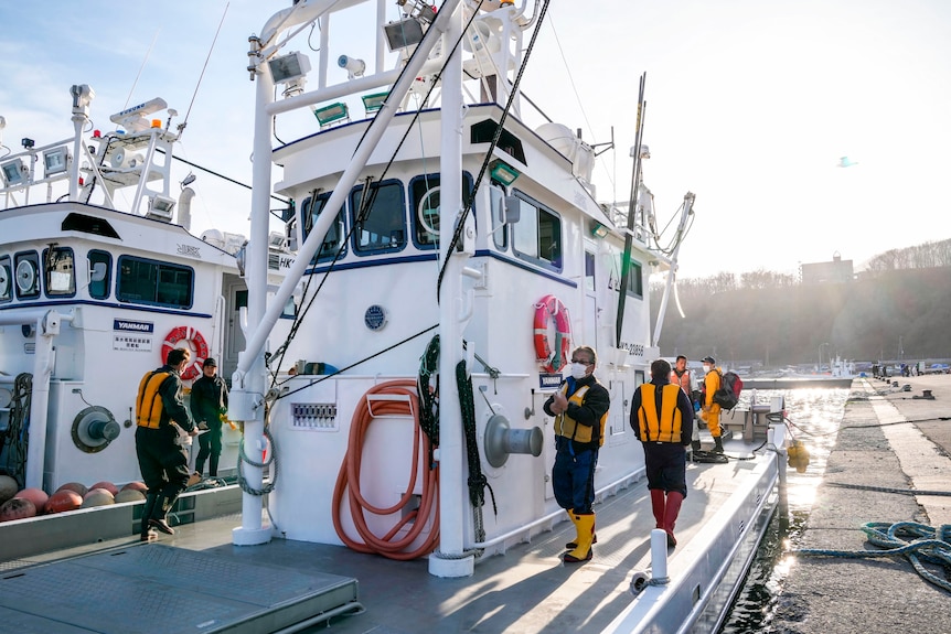 The crew members of a fishing boat stand on its deck, dressed in high-jackets and high boots.