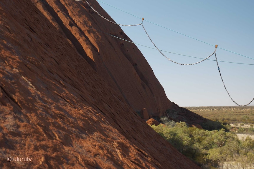 The chain is seen being removed from Uluru. It is attached to a pulley system and the rock is visible behind.
