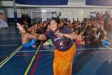 Young woman wears blue tee and orange ie lava lava. Arms outstretched as she dances the Samoan siva. Students sit behind her. 