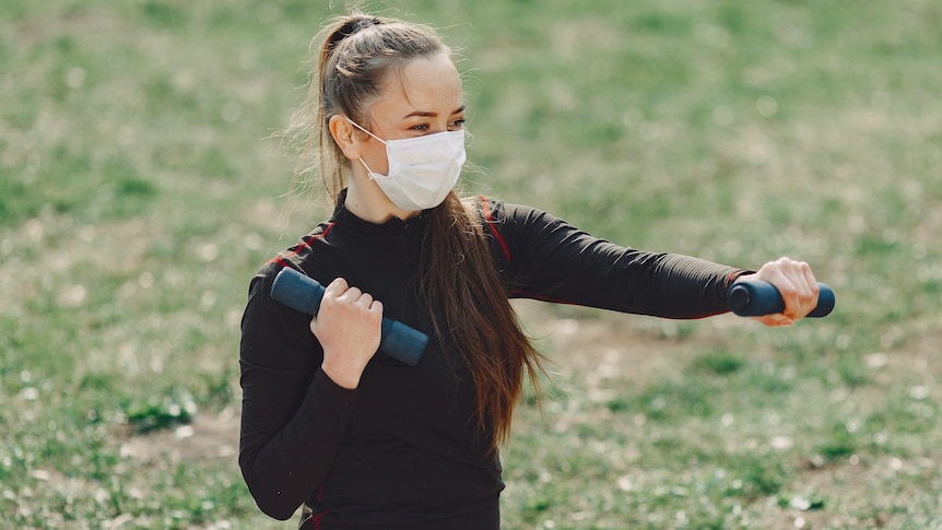 Woman wears a black top and leggings and holds dumbbells and wears a mask, for story about exercising while wearing mask..