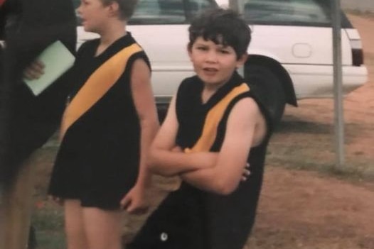 A young boy with crossed arms wearing footy kit looks at the camera
