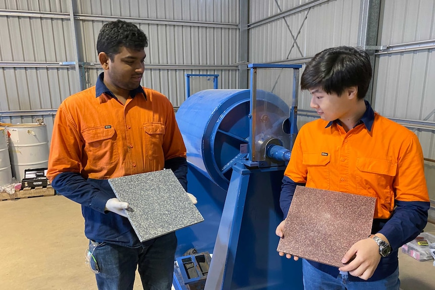 Two men in hi-vis orange shirts hold a tile each in a shed