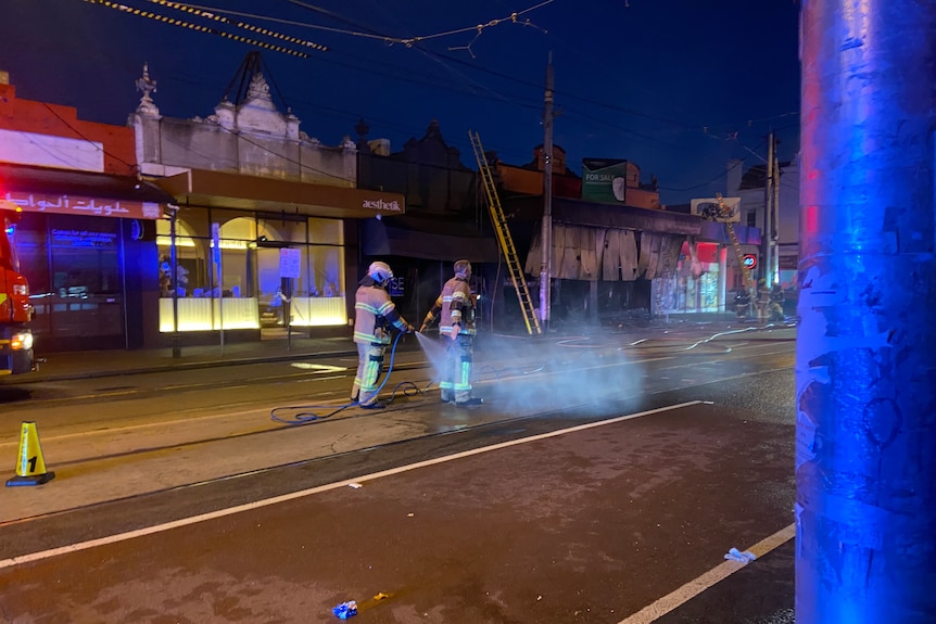 A firefighter is hosed down with water by another firefighter in the middle of tram tracks on a dark road.