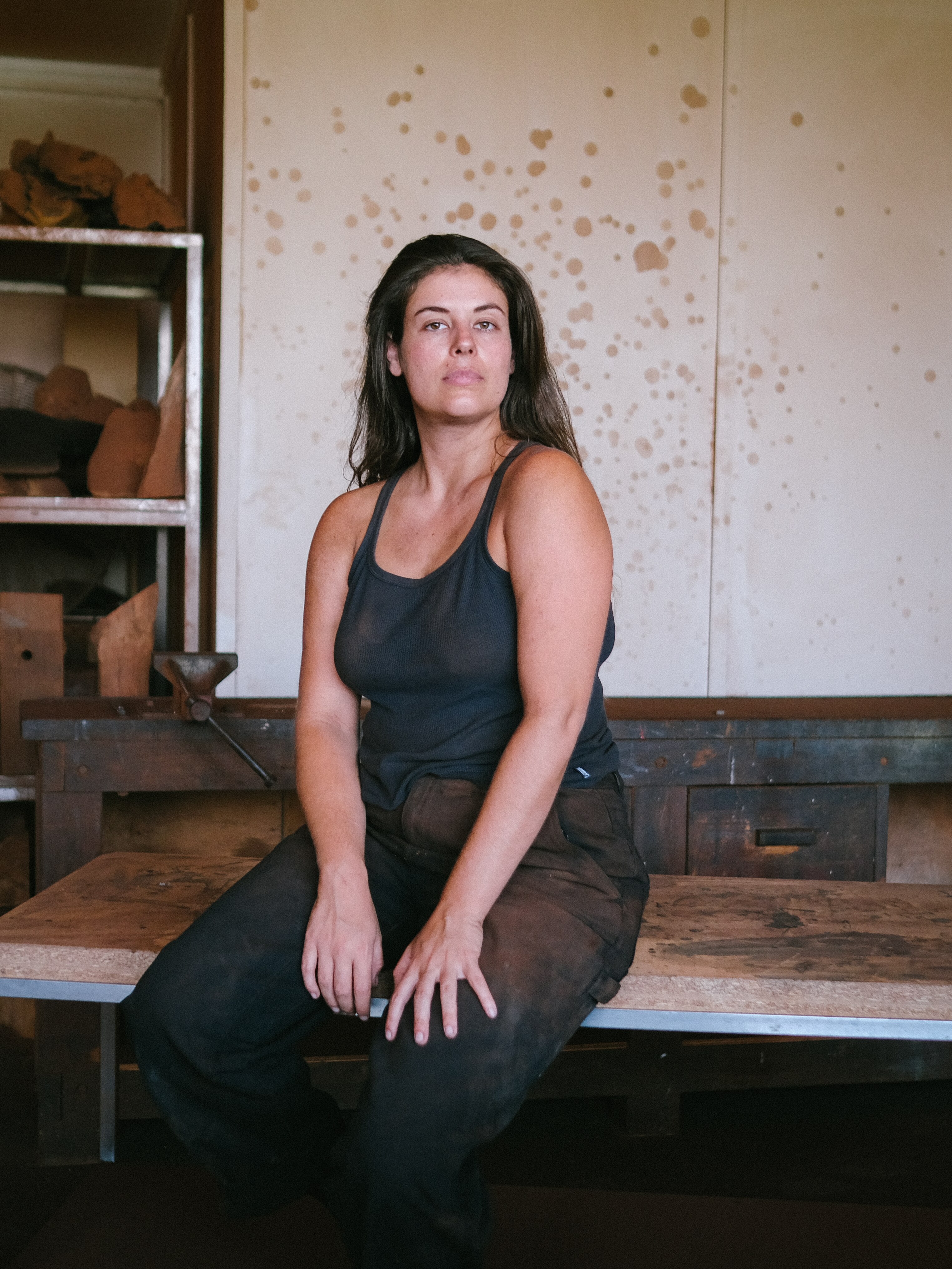 Portrait of artist Olive Gill-Hille, wearing black, sitting on a bench in her studio facing the camera 