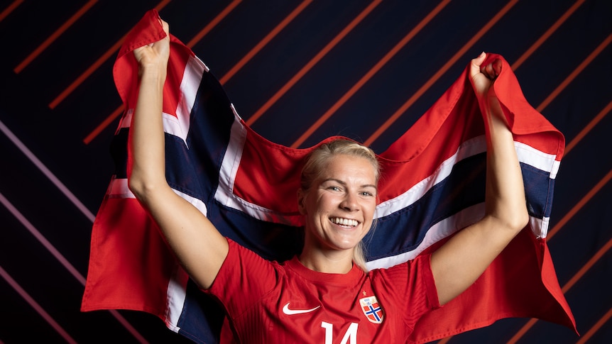 A Norwegian football star dressed in team colours smiles as she holds a Norwegian flag behind her back. 