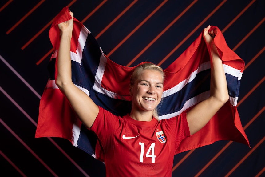 A Norwegian football star dressed in team colours smiles as she holds a Norwegian flag behind her back. 