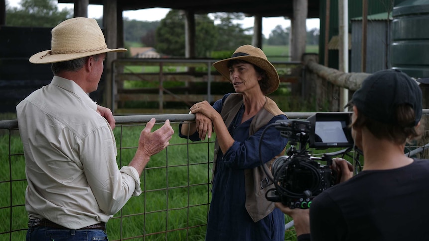 A woman in a wide-brimmed hat talks to a farmer as they are filmed by a cameraman