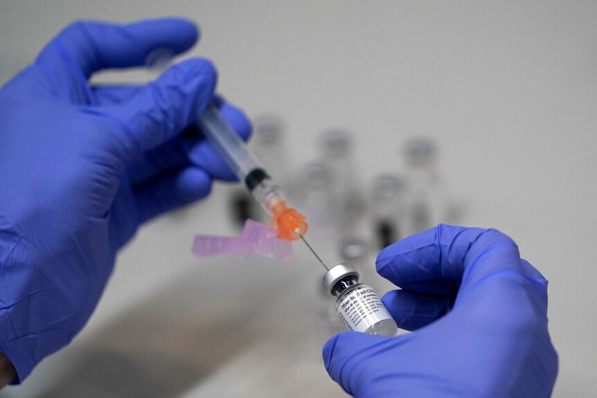 a person wearing blue gloves holds a needle and a vial of covid vaccine in their hands