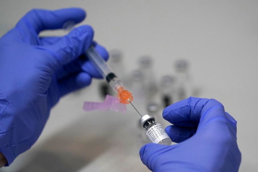 a person wearing blue gloves holds a needle and a vial of covid vaccine in their hands