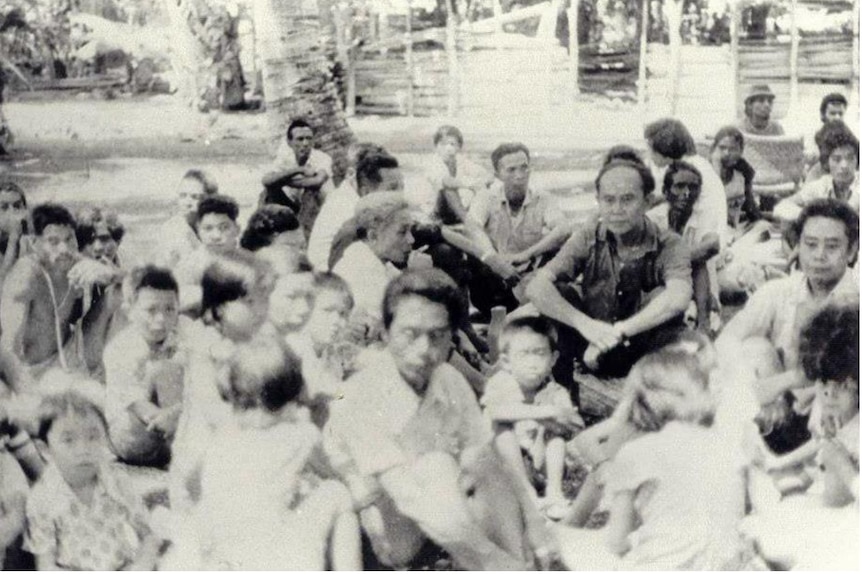 Grainy, blurry black and white photo of group of people of varying ages sitting on the ground.