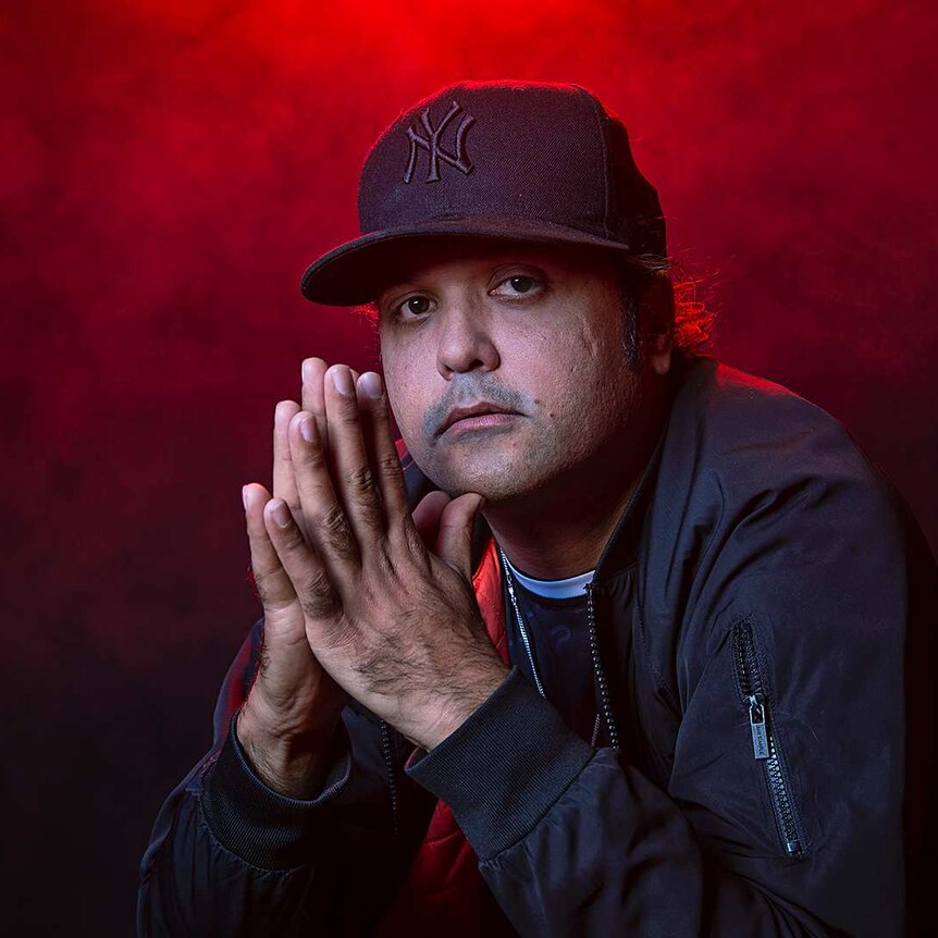 Colour portrait of rapper Birdz with palms clasped together in front of black background and some red light.