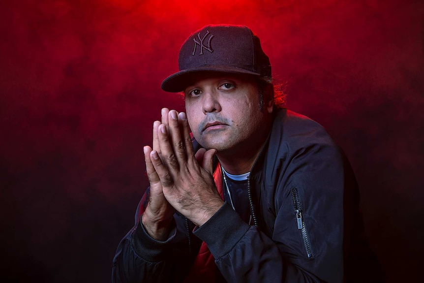 Colour portrait of rapper Birdz with palms clasped together in front of black background and some red light.