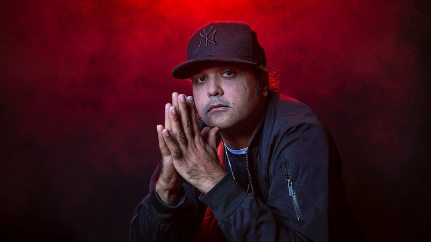 Colour portrait of rapper Birdz with palms clasped together in front of black background and some red light.