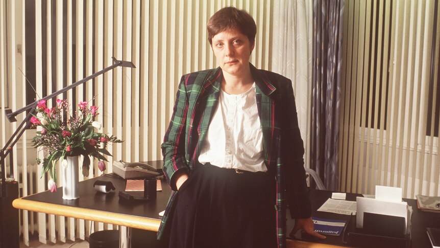 A young Angela Merkel with short hair dressed in a check 80's jacket leans against a desk looking directly at the camera 